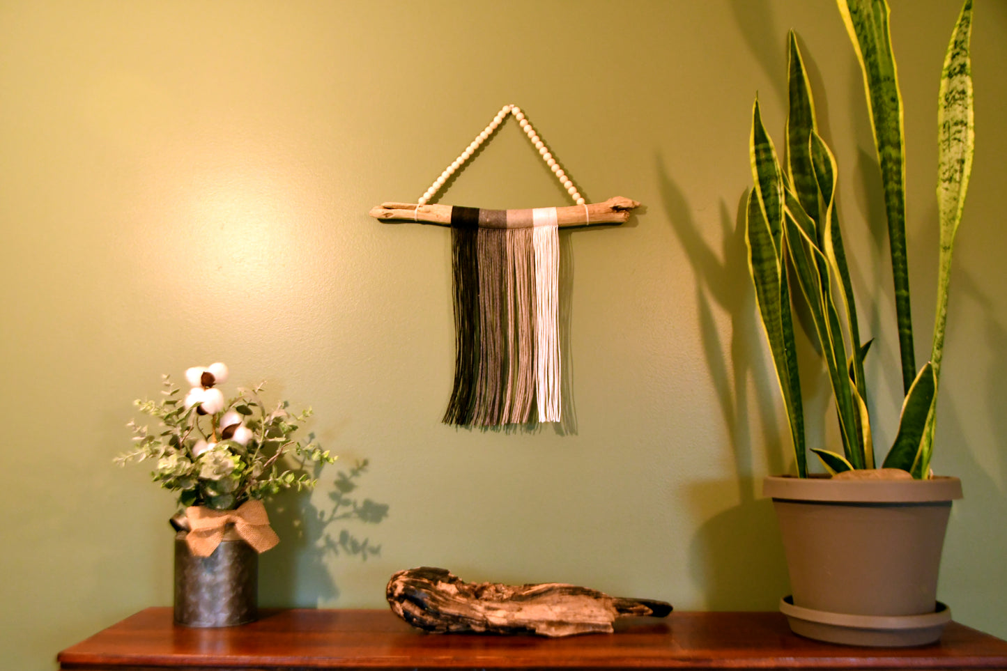 Woven wall art piece on a green wall with a plant and vase on a wooden surface.
